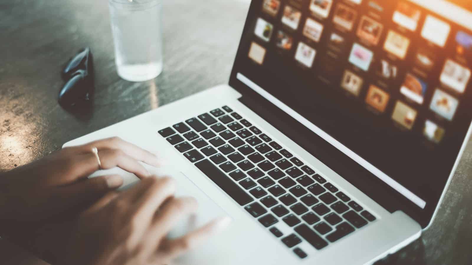 A person is typing on a laptop with a display showing various image thumbnails. The scene is well-lit with natural light. Sunglasses are placed on the table next to the laptop.