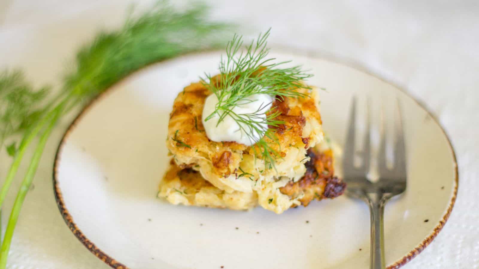 A serving of potato and herb fritter topped with a dollop of sour cream and fresh dill is on a white plate. A metal fork and a sprig of dill are placed beside the fritter. The background is a textured white surface.