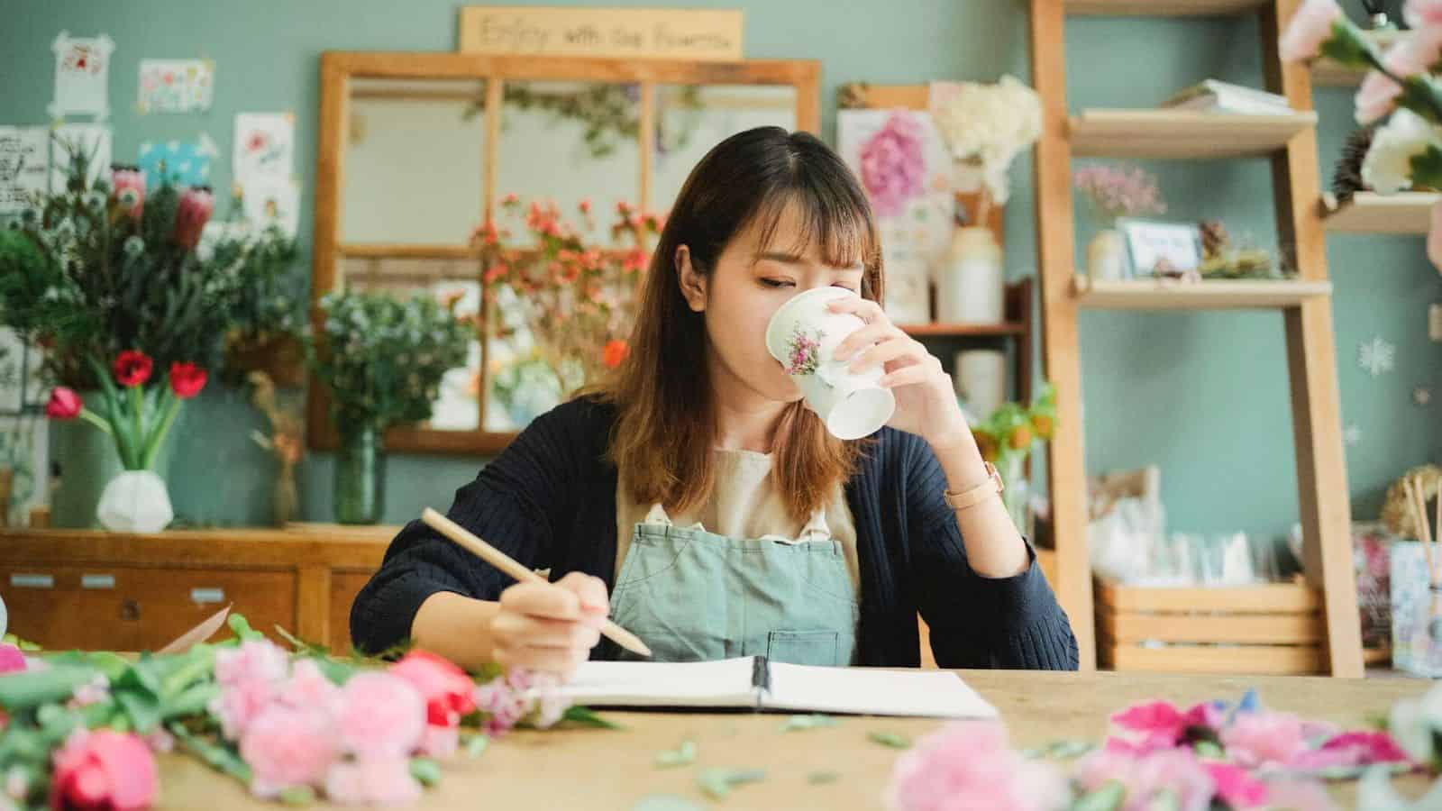A woman wearing an apron sits at a table in a flower shop. She is drinking from a floral mug while writing in a notebook. Various flowers and leaves are scattered across the table, and shelves with plants are visible in the background.