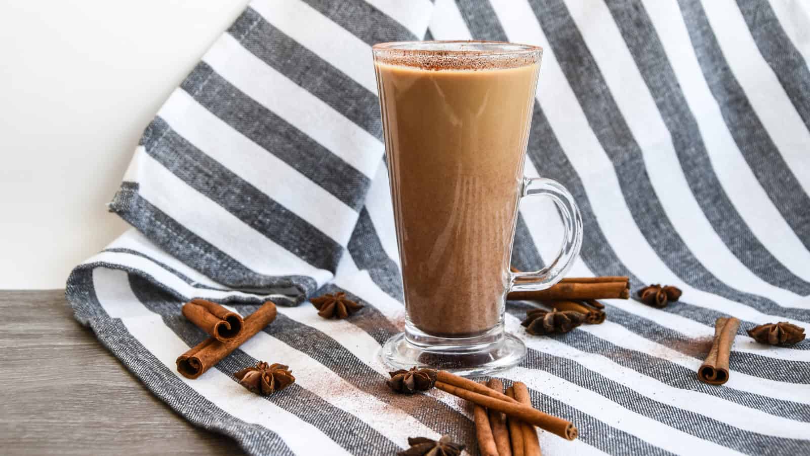 A glass mug of chocolate milk stands on a gray and white striped cloth. Cinnamon sticks and star anise are scattered around the mug. The scene is set on a wooden surface.