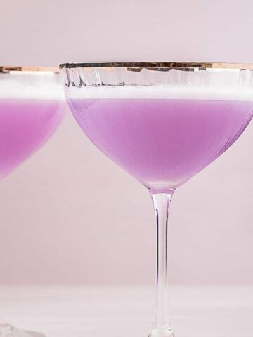 Two coupe glasses filled with a lavender-colored gin cocktail are placed on a white surface. A bottle of purple gin stands nearby. In the background, there's a vase with dried flowers.
