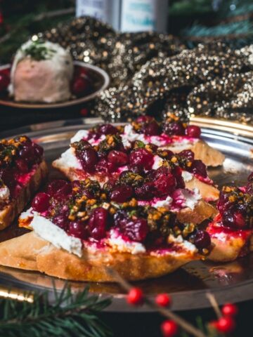A platter of bruschetta topped with cheese, cherries, and herbs on a gold-edged metal tray. The background features a festive setting with a sparkling cloth, greenery, and a glass of red beverage.