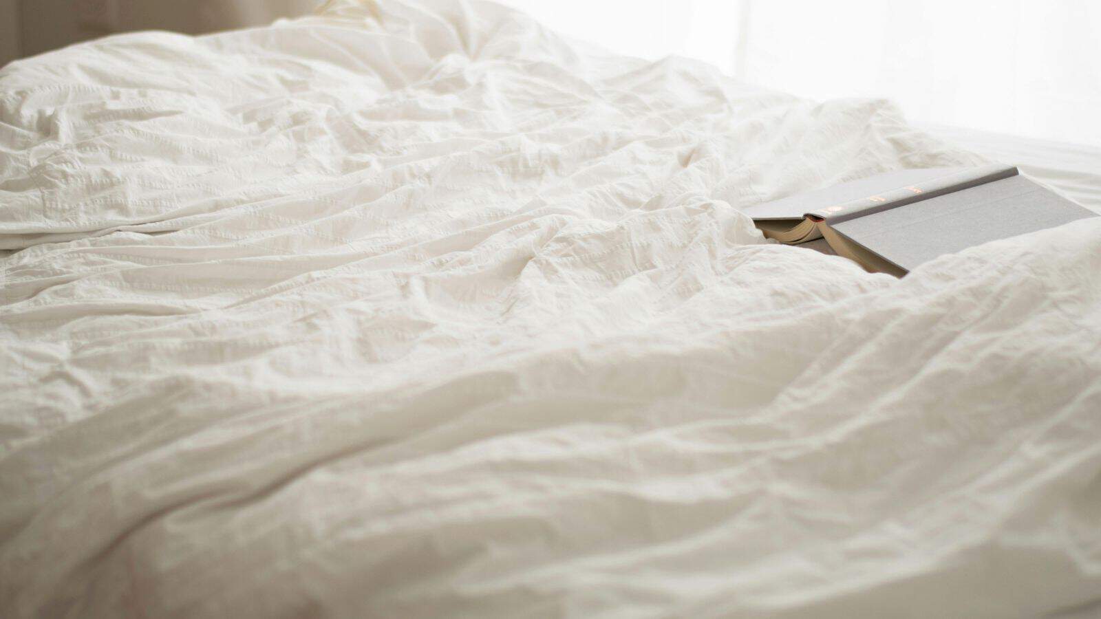 A neatly made bed with a white textured duvet covers most of the image. Natural light filters in from a window, and a closed book with a light gray cover is placed on the right side of the bed.