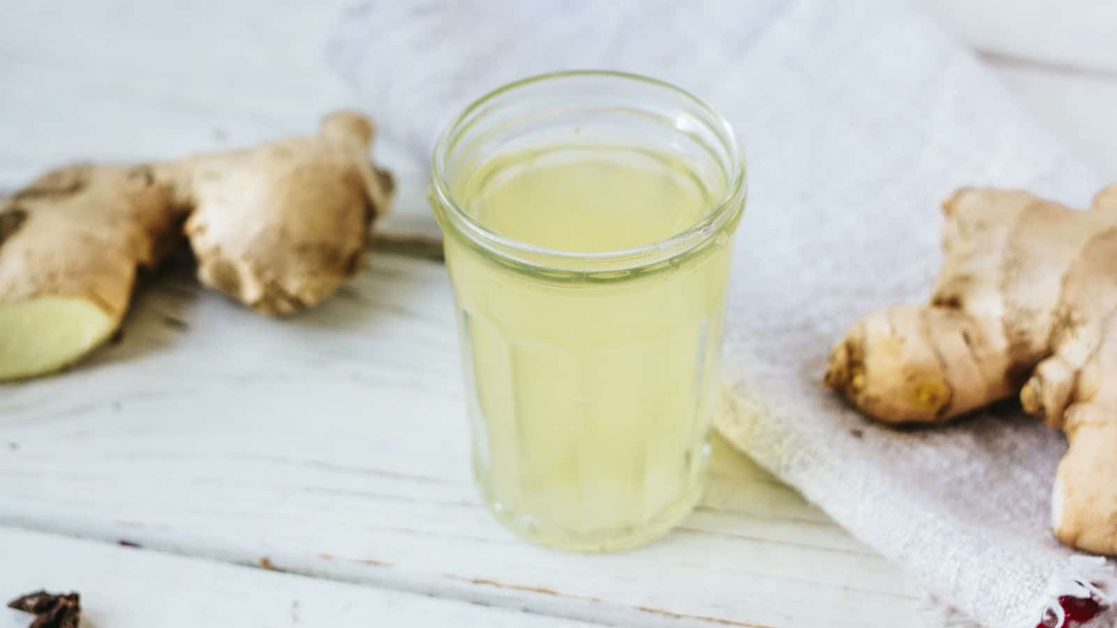 A glass of ginger juice is placed on a white wooden surface. Fresh ginger roots are nearby, along with a light-colored cloth.