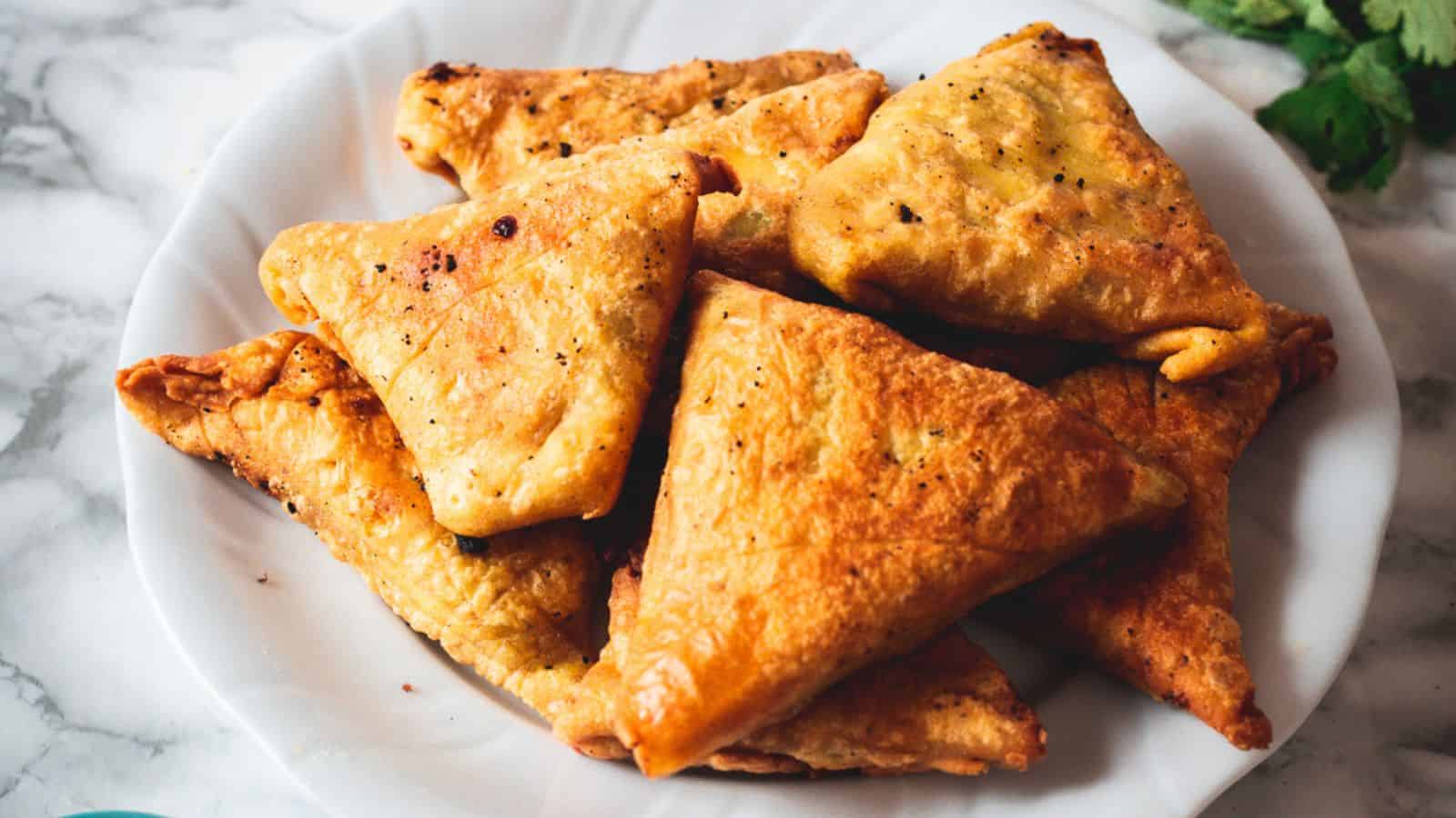A plate of golden-brown, triangular pastries, likely samosas, is arranged on a white dish. The pastries are seasoned with visible spices and resting on a marble surface. Fresh coriander leaves are partially visible in the background.