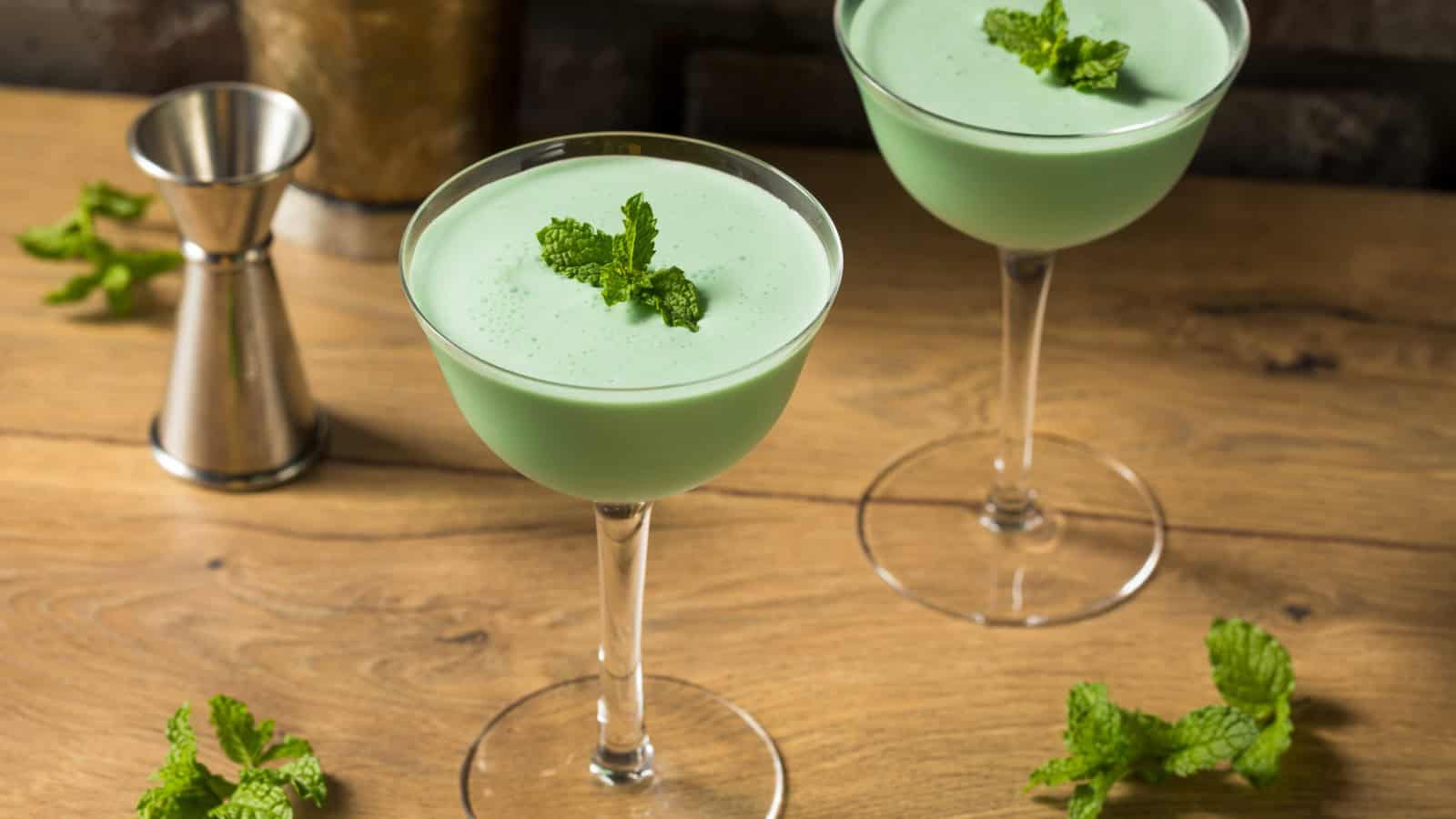 Two cocktail glasses filled with a green creamy drink are placed on a wooden surface. Each glass is garnished with a mint sprig. A metal jigger and scattered mint leaves are in the background.