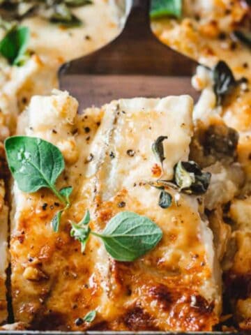 A close-up of a casserole dish with a baked lasagna. The golden-brown cheese topping is garnished with fresh oregano leaves. One portion of lasagna is cut out, revealing layers of pasta, sauce, and cheese.