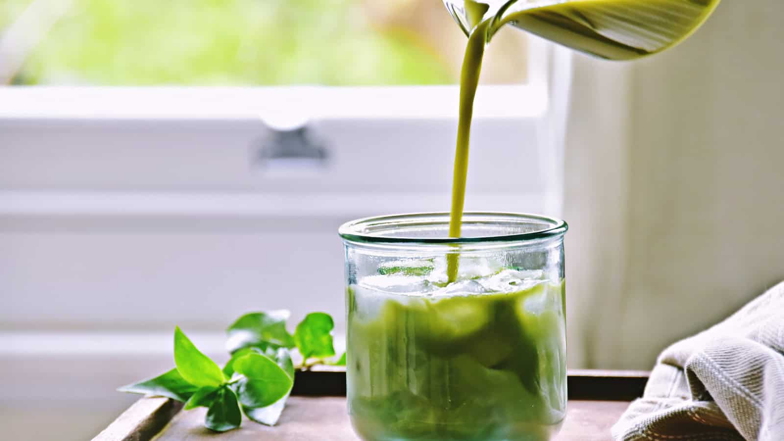 A glass filled with ice sits on a wooden tray. A green liquid is being poured into the glass from a pitcher. A leafy plant is in the background, and a white window is partially visible.