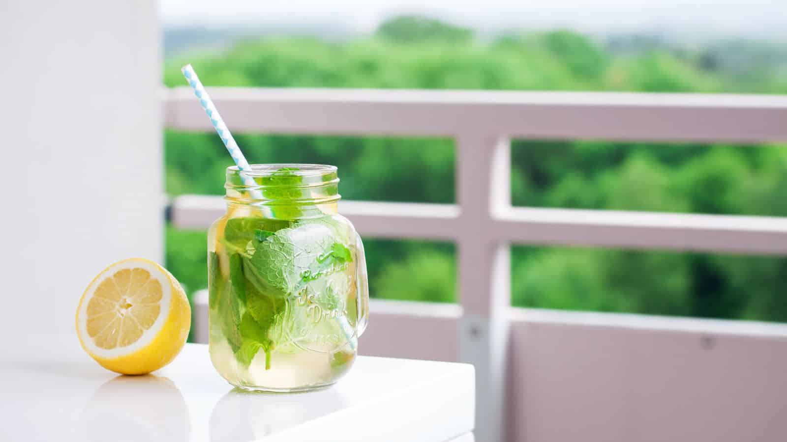 A mason jar filled with a minty beverage and a striped straw sits on a white surface. A halved lemon is placed beside the jar. The background features a blurred view of greenery and a pink railing.