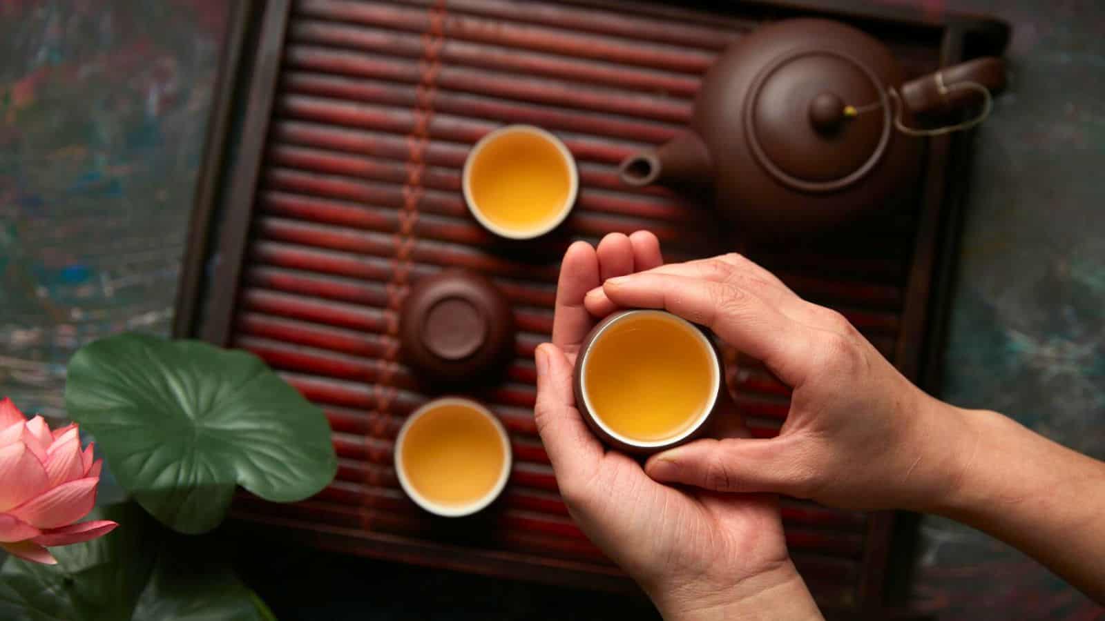 A pair of hands holds a small cup filled with tea. The cup is on a wooden tray with two other tea cups, a teapot, a green leaf, and a pink lotus flower. The background is a mix of textures and colors.