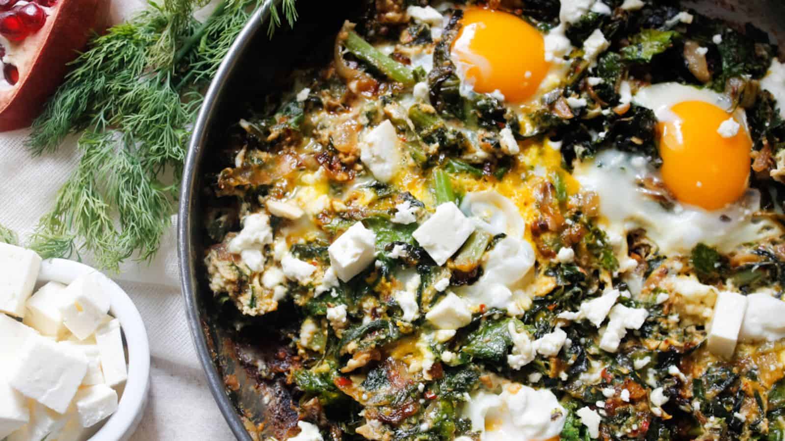A close-up of a skillet containing a cooked dish of leafy greens, two sunny-side-up eggs, and crumbled feta cheese. Fresh dill and a bowl of extra feta are on the side. A portion of a pomegranate is partially visible in the corner.
