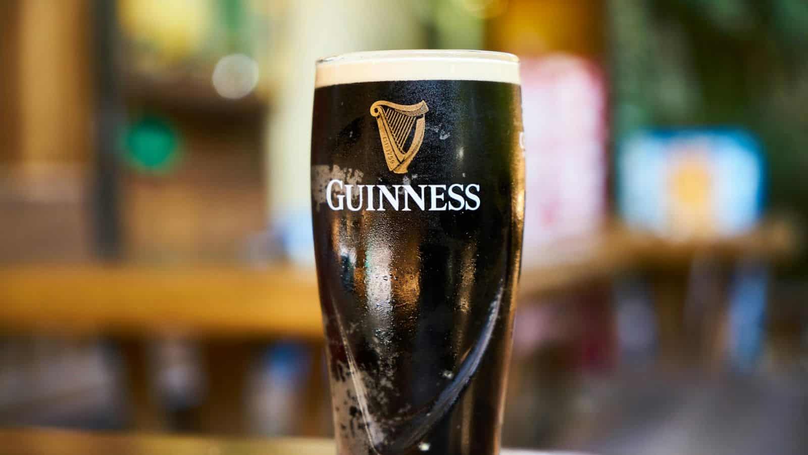 A glass of dark Guinness beer with a creamy head is placed on a wooden table, in an indoor setting with blurred background elements. The Guinness logo is visible on the glass.