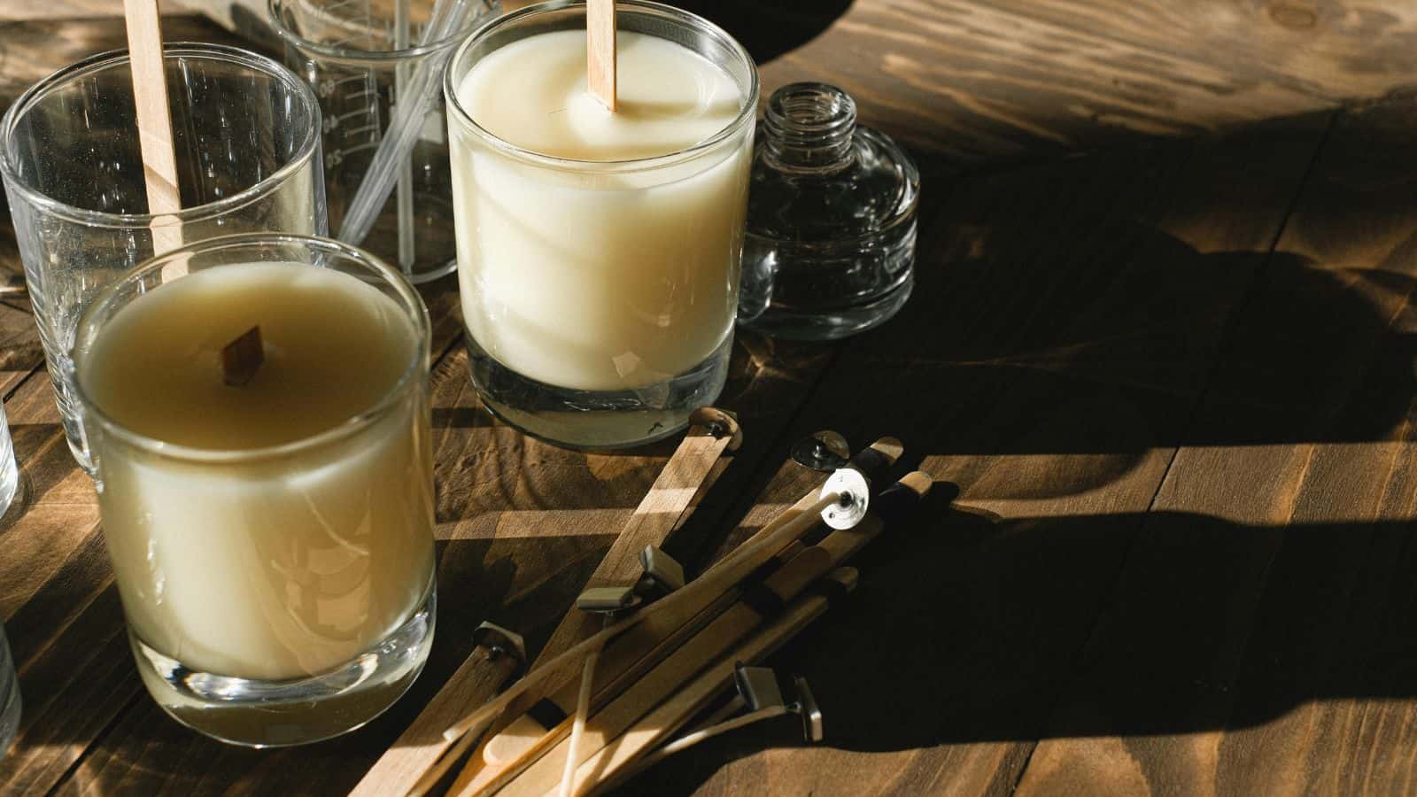 Glass candles with light beige wax and wooden wicks rest on a wooden table. Nearby are an empty glass bottle and scattered wooden candle wicks with metal bases. Light streaming from a window casts shadows across the surface.