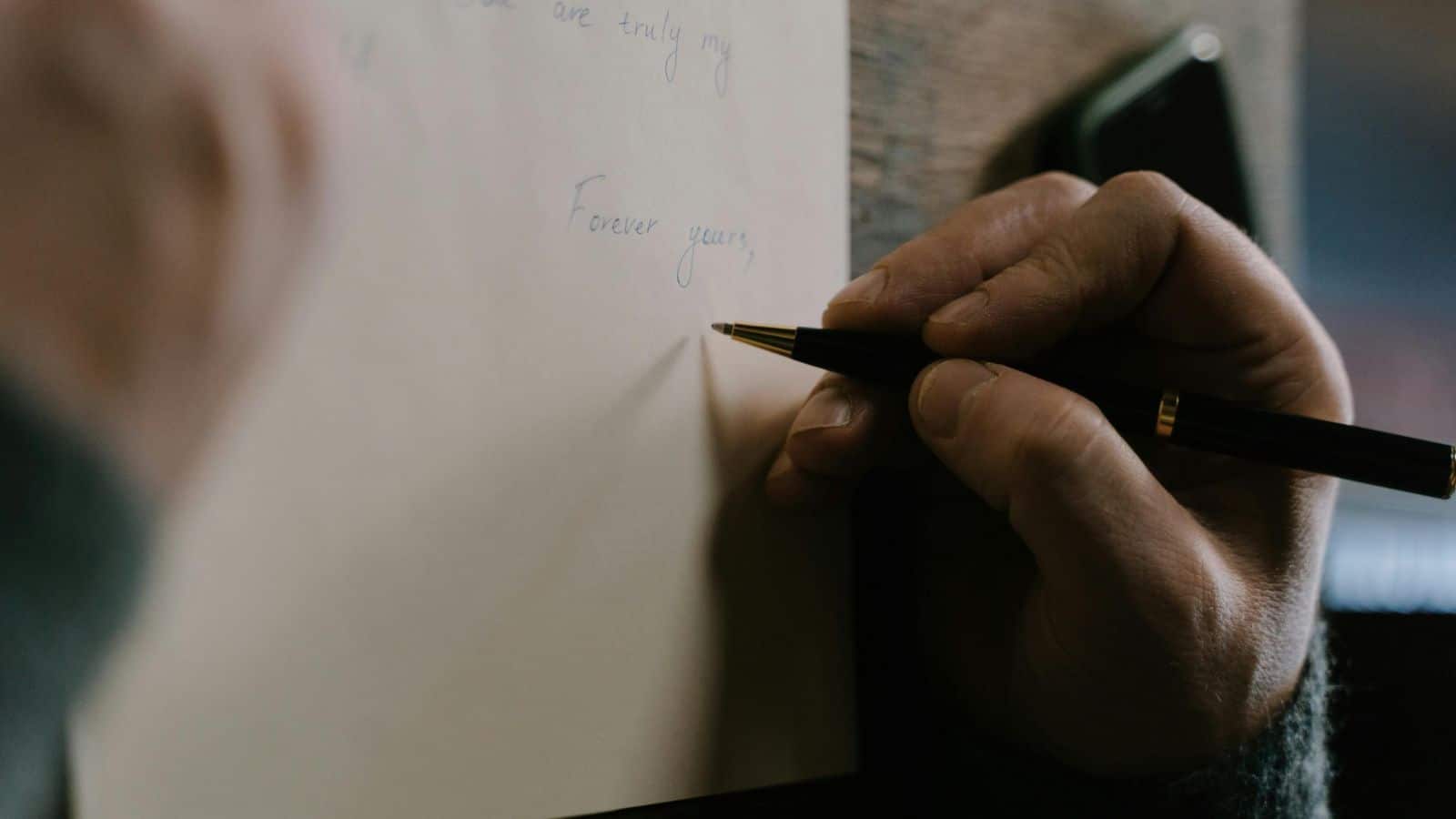 A close-up of a hand writing on a piece of paper with a black pen. The paper contains part of a handwritten message, with the words "Forever yours" visible. A smartphone lies nearby on the wooden surface.