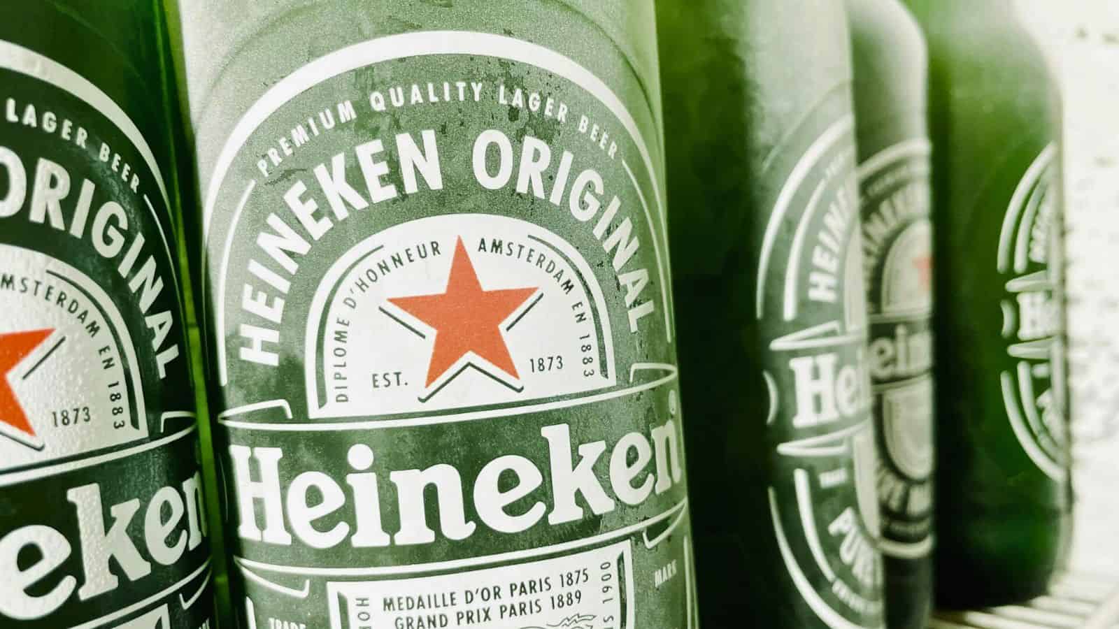 Close-up of several green Heineken beer bottles aligned in a row. The labels display "Heineken Original" with a red star logo, emphasizing its Amsterdam and lager origins.