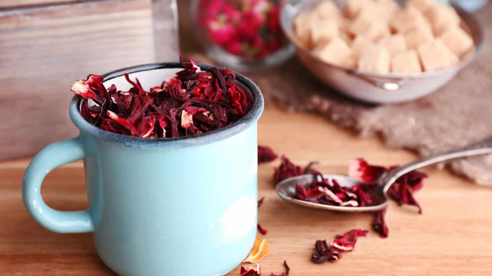 A blue mug filled with dried hibiscus petals is on a wooden surface. A spoon with petals and a bowl of sugar cubes are in the background, along with a blurred out container with more petals.