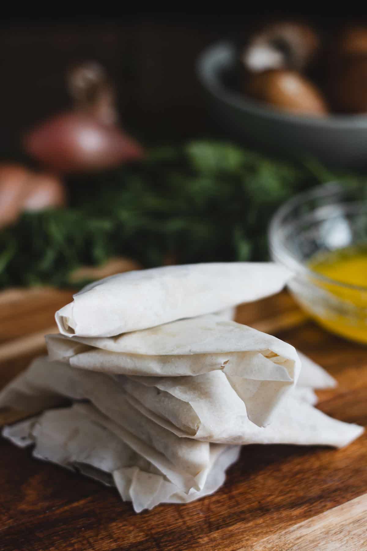 A stack of wrapped, uncooked phyllo pastry rolls awaits transformation into savoury pastries on a wooden surface. Beside it, a glass bowl brims with melted butter against a blurred backdrop of fresh herbs and vegetables.