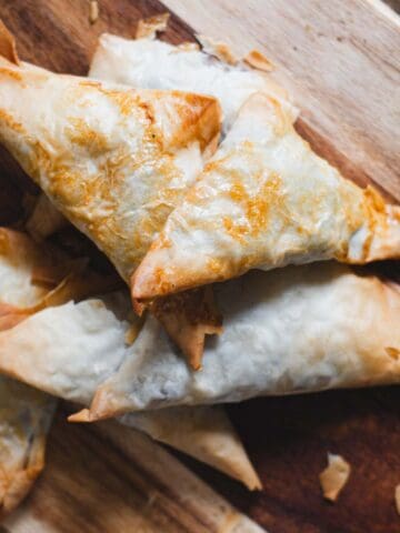 A wooden board with savoury, baked triangular pastries next to fresh parsley, shallots, and a bowl of mushrooms on a rustic table.