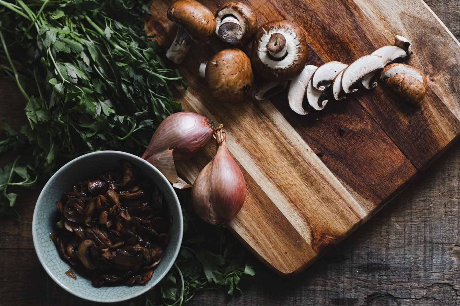 A wooden cutting board with whole and sliced mushrooms, surrounded by herbs, sets the stage for creating savory pastries. Nearby, a bowl contains cooked mushrooms ready for filling, while two shallots are poised beside the board on a rustic wooden surface.