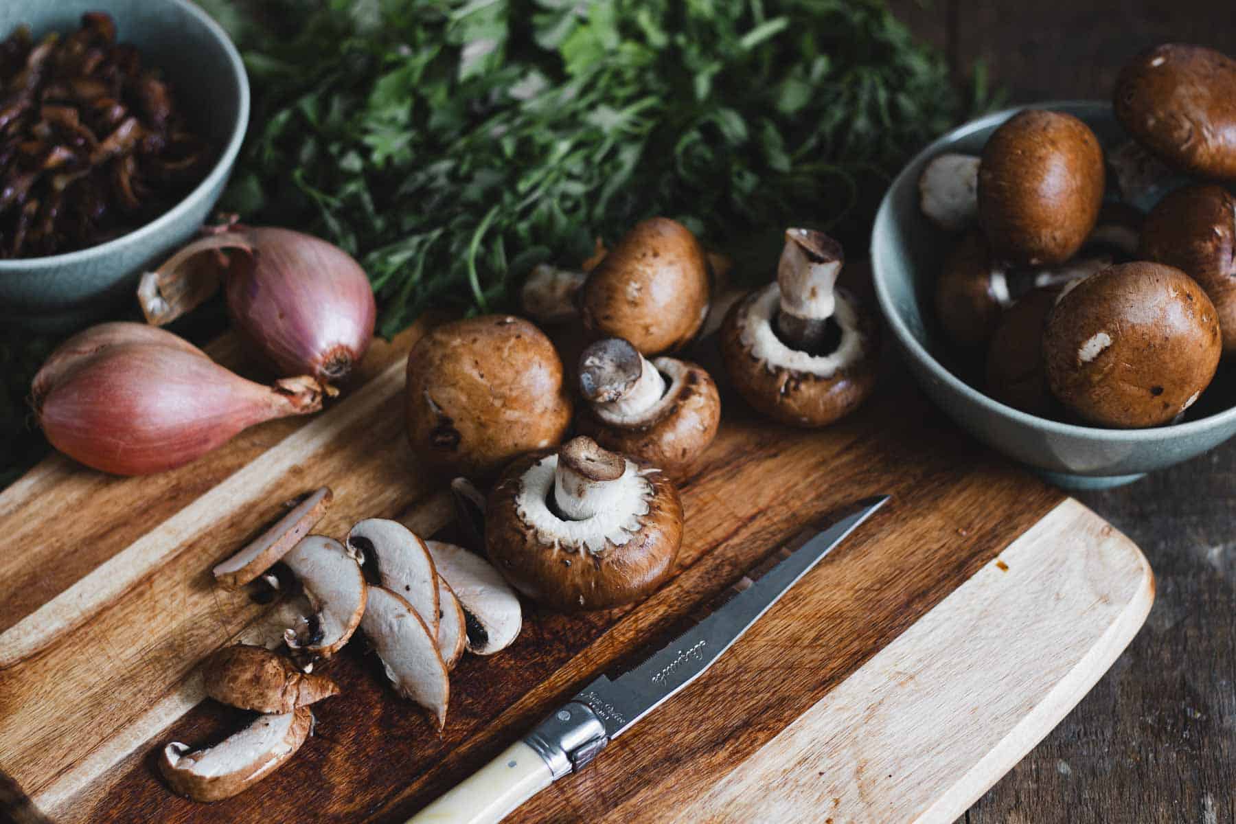 A wooden cutting board with whole and sliced mushrooms, a knife, and shallots sets the stage for crafting savory pastries. Bowls brimming with additional mushrooms and herbs linger in the background, promising layers of flavor.