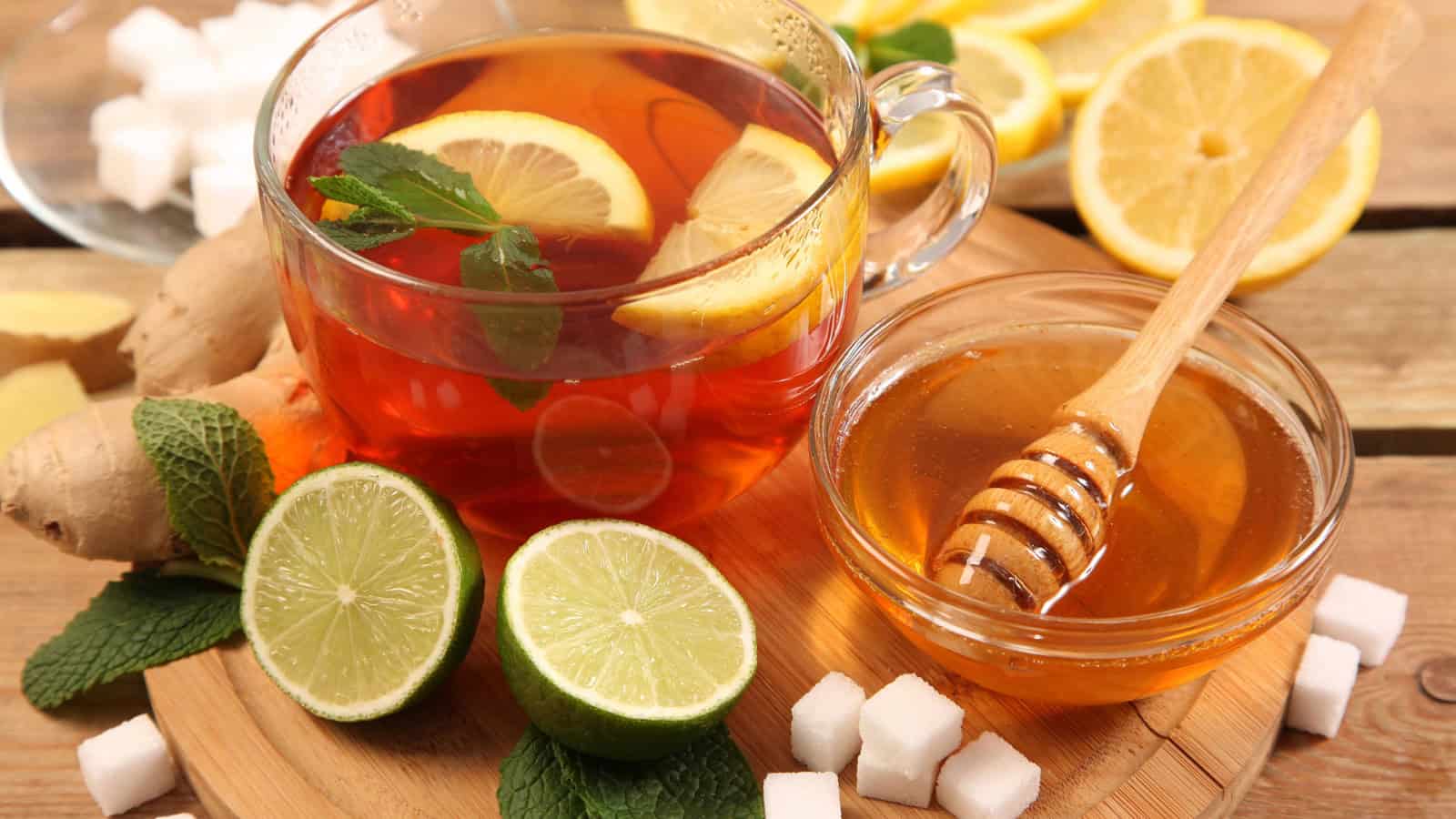 A glass cup of tea with lemon slices and mint leaves sits on a wooden tray alongside a small bowl of honey with a honey dipper. Surrounding the tray are ginger, lime halves, lemon slices, mint leaves, and sugar cubes.