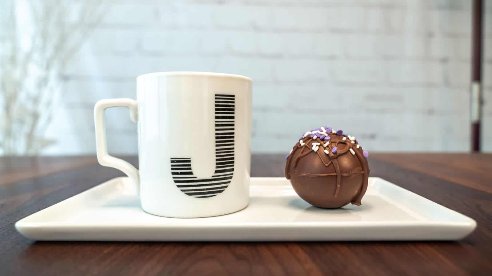 A white mug with a large black letter "J" sits on a rectangular white tray alongside a chocolate sphere garnished with white and purple sprinkles. The background is a blurred white brick wall.