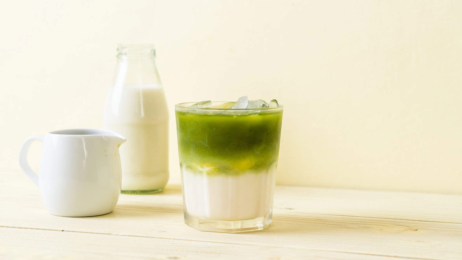 A glass of iced matcha latte, reminiscent of underrated Starbucks flavors, showcases distinct green and white layers on a wooden surface. To the left, a small white pitcher and a glass bottle brimming with milk complement the light cream backdrop.