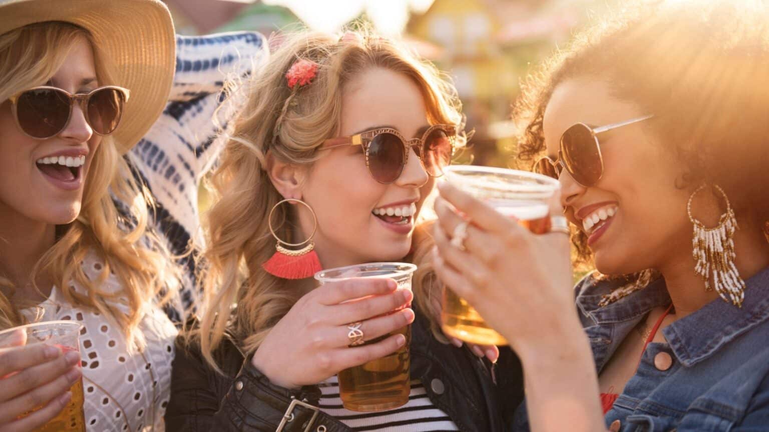 Three women are enjoying drinks outdoors in sunny weather. They are wearing sunglasses and smiling, with one wearing a sunhat and the others wearing large earrings. They are holding plastic cups filled with beverages.