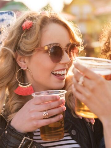 Three women are enjoying drinks outdoors in sunny weather. They are wearing sunglasses and smiling, with one wearing a sunhat and the others wearing large earrings. They are holding plastic cups filled with beverages.