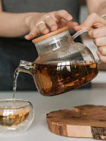 A person pours tea from a glass teapot into a double-walled glass cup. The teapot has a wooden lid, and the cup is placed on a white surface next to a round wooden coaster. An open magazine is partially visible.