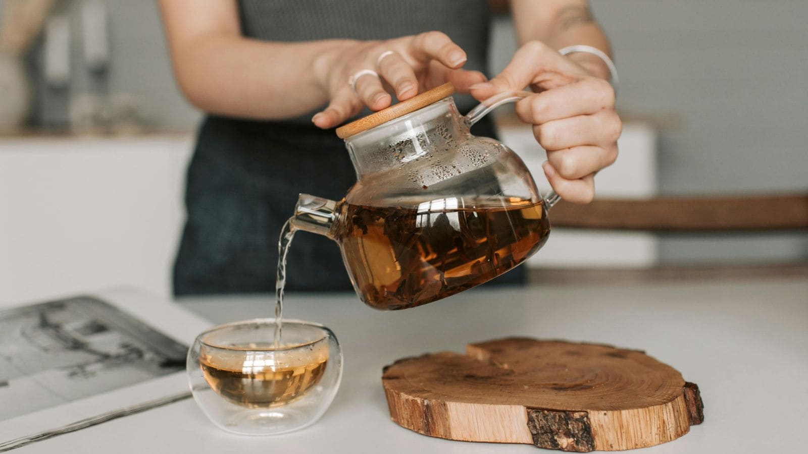A person pours tea from a glass teapot into a double-walled glass cup. The teapot has a wooden lid, and the cup is placed on a white surface next to a round wooden coaster. An open magazine is partially visible.