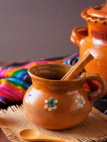 A rustic setup featuring a clay pot with a lid, a smaller clay mug with floral designs containing a cinnamon stick, a colorful woven cloth, chunks of unrefined sugar on a burlap square, a wooden spoon, and a small bowl of coffee beans.
