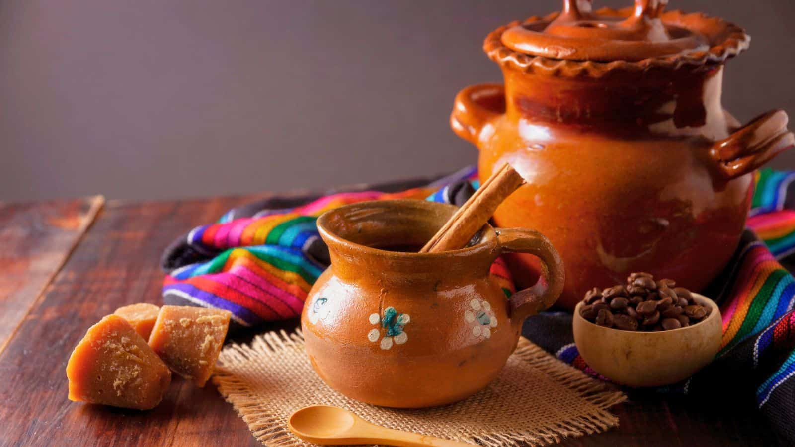 A rustic setup featuring a clay pot with a lid, a smaller clay mug with floral designs containing a cinnamon stick, a colorful woven cloth, chunks of unrefined sugar on a burlap square, a wooden spoon, and a small bowl of coffee beans.