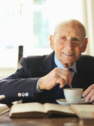 An elderly man with glasses sits at a table, smiling and stirring a cup of tea or coffee. Several open books are spread out in front of him, with a window and curtain in the background.