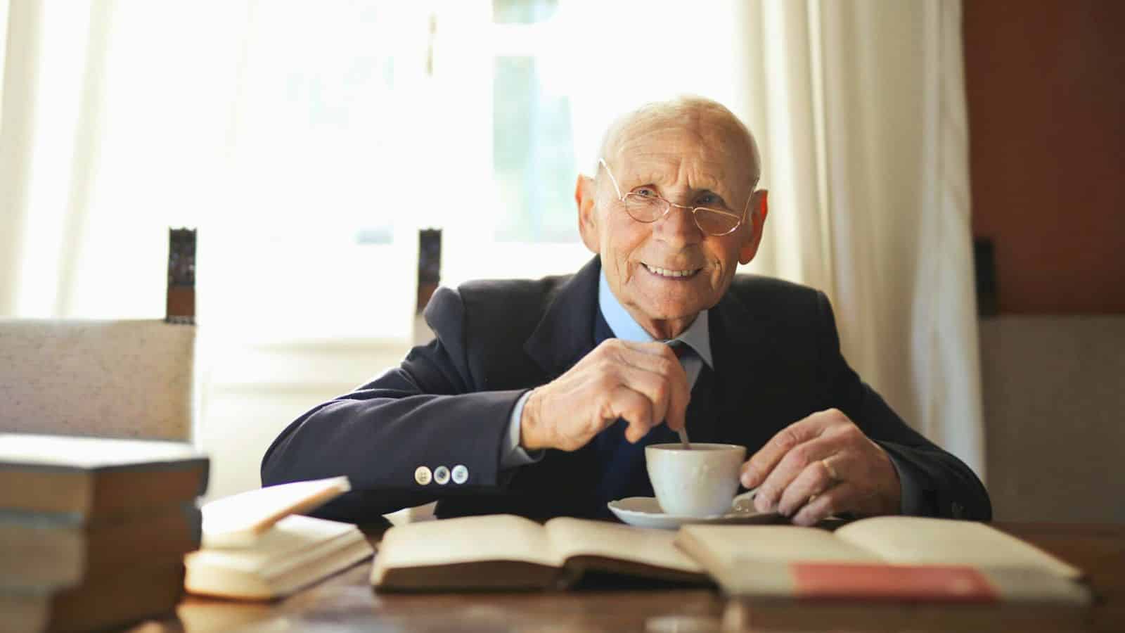 An elderly man with glasses sits at a table, smiling and stirring a cup of tea or coffee. Several open books are spread out in front of him, with a window and curtain in the background.
