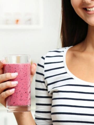 A woman is holding a glass of pink smoothie in a kitchen. She is wearing a black and white striped shirt and smiling. The background is slightly blurred, showing kitchen elements like jars and a counter.