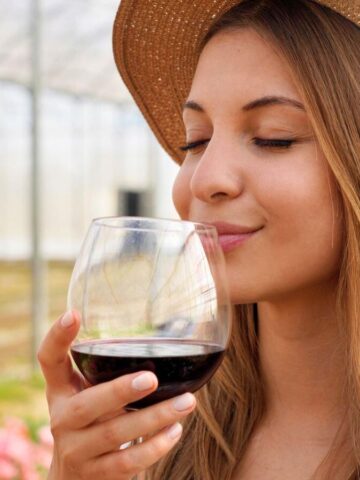 A woman with long hair is wearing a straw hat and holding a glass of red wine close to her nose. She is standing in a greenhouse filled with plants and flowers. The background is blurred, creating a peaceful atmosphere.