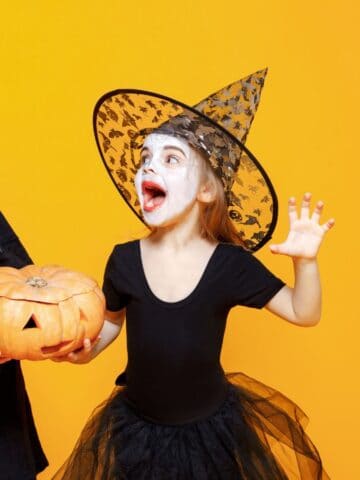 A young boy in a black costume with a green face paint holds a carved pumpkin. Next to him, a young girl wears a witch's hat and black outfit, with her face painted white and red eyeshadow. Both children are set against an orange background.