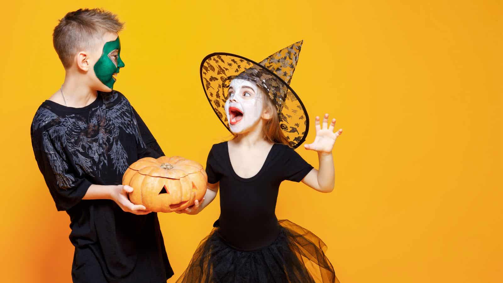 A young boy in a black costume with a green face paint holds a carved pumpkin. Next to him, a young girl wears a witch's hat and black outfit, with her face painted white and red eyeshadow. Both children are set against an orange background.