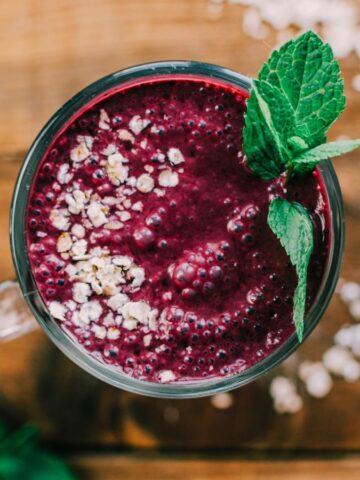 A top-down view of a purple smoothie in a glass, garnished with oats and a sprig of mint. The smoothie is placed on a wooden surface with a few oats scattered around.