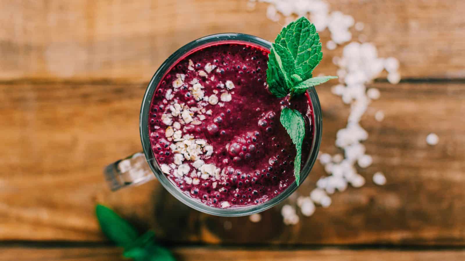 A top-down view of a purple smoothie in a glass, garnished with oats and a sprig of mint. The smoothie is placed on a wooden surface with a few oats scattered around.