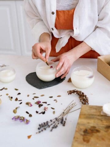 A person is making candles on a white kitchen table. There are three glass candles, dried flowers, herbs, and wooden discs on the table. A box with more dried materials and a drink are nearby. A stove can be seen in the background.