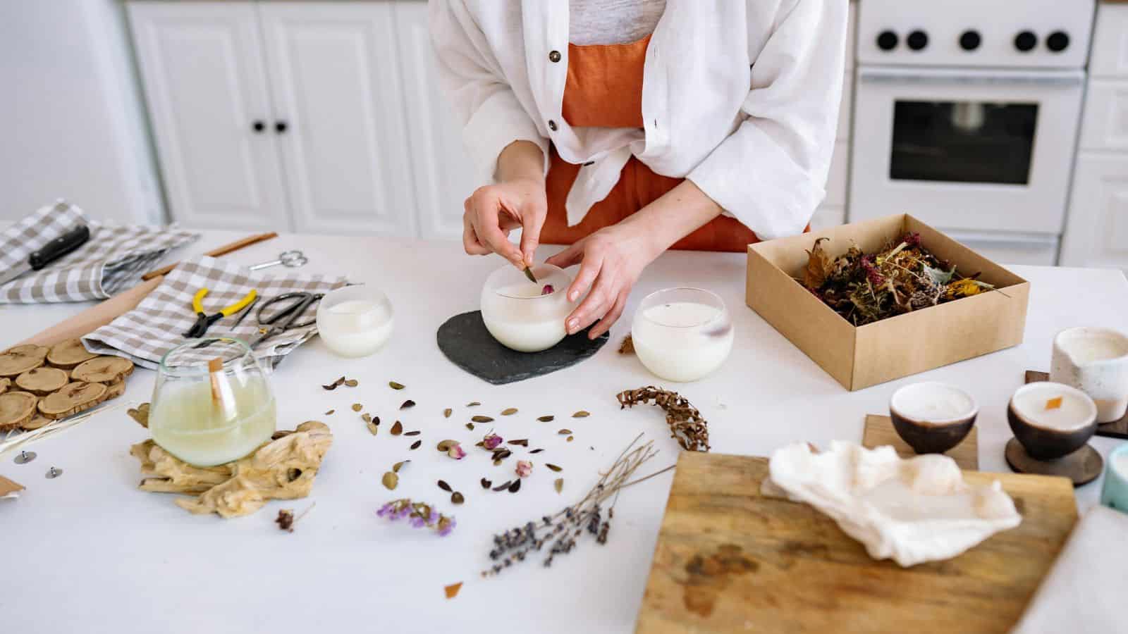 A person is making candles on a white kitchen table. There are three glass candles, dried flowers, herbs, and wooden discs on the table. A box with more dried materials and a drink are nearby. A stove can be seen in the background.