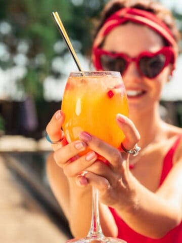 A person in a red swimsuit, holding a vibrant orange cocktail with a straw, stands near a swimming pool. They are wearing heart-shaped sunglasses and a red headband. The background shows blurred trees and poolside umbrellas.