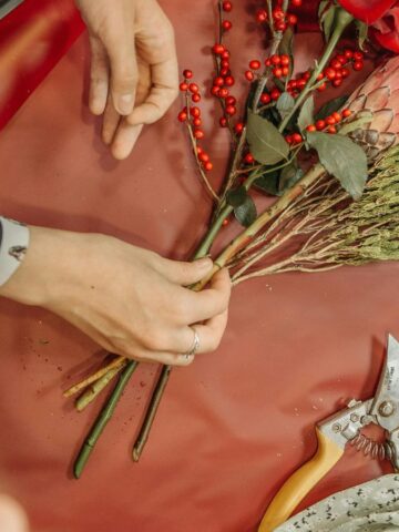 Three people arranging a floral bouquet on a red sheet. The bouquet includes red berries, green foliage, and a pink protea. Scissors are nearby on the table. Hands are adjusting the flowers and wrapping paper.