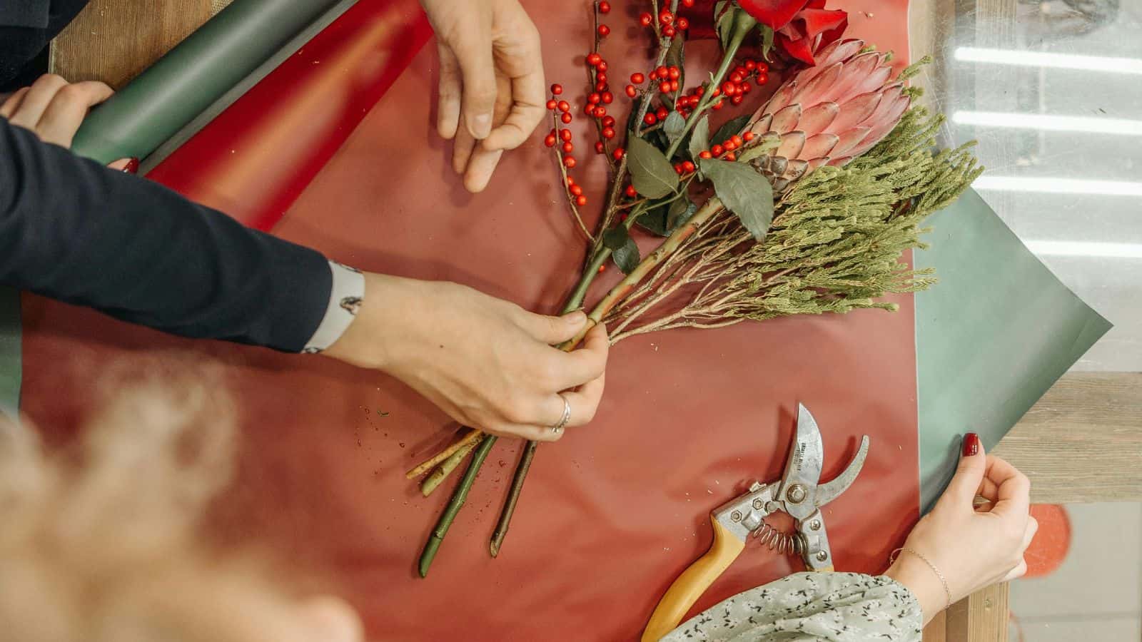 Three people arranging a floral bouquet on a red sheet. The bouquet includes red berries, green foliage, and a pink protea. Scissors are nearby on the table. Hands are adjusting the flowers and wrapping paper.