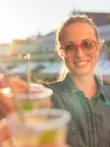 A person wearing red sunglasses and a denim shirt stands outdoors at a sunlit market, holding a drink with a smile. The background shows blurred market stalls and people.