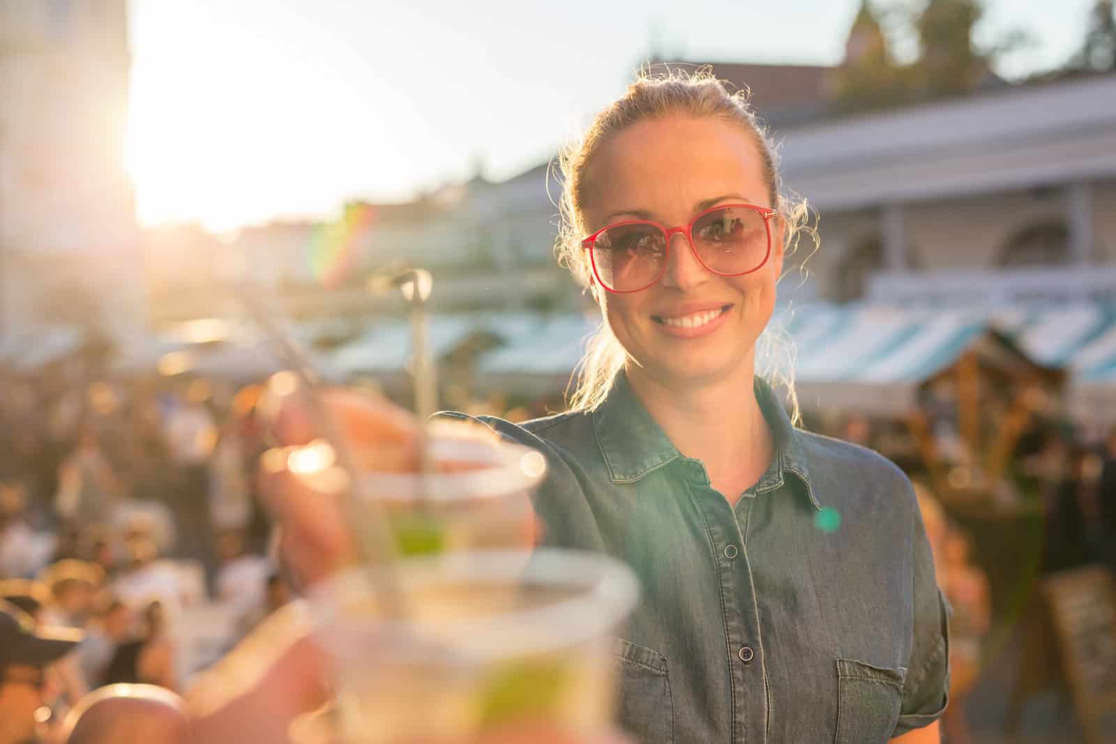 A person wearing red sunglasses and a denim shirt stands outdoors at a sunlit market, holding a drink with a smile. The background shows blurred market stalls and people.