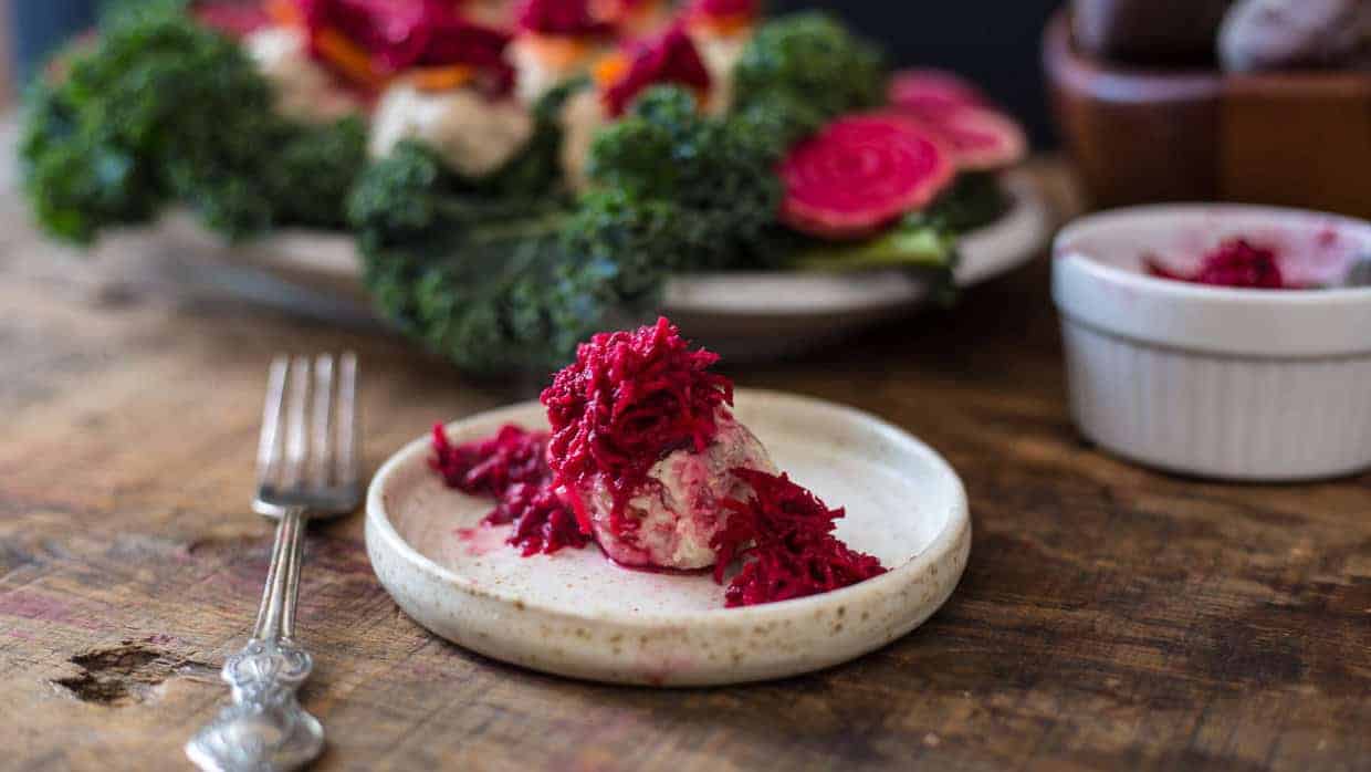A small plate with a scoop of food topped with shredded beetroot, accompanied by a fork on a wooden table. In the background, a larger dish with leafy greens and more beetroots is visible.