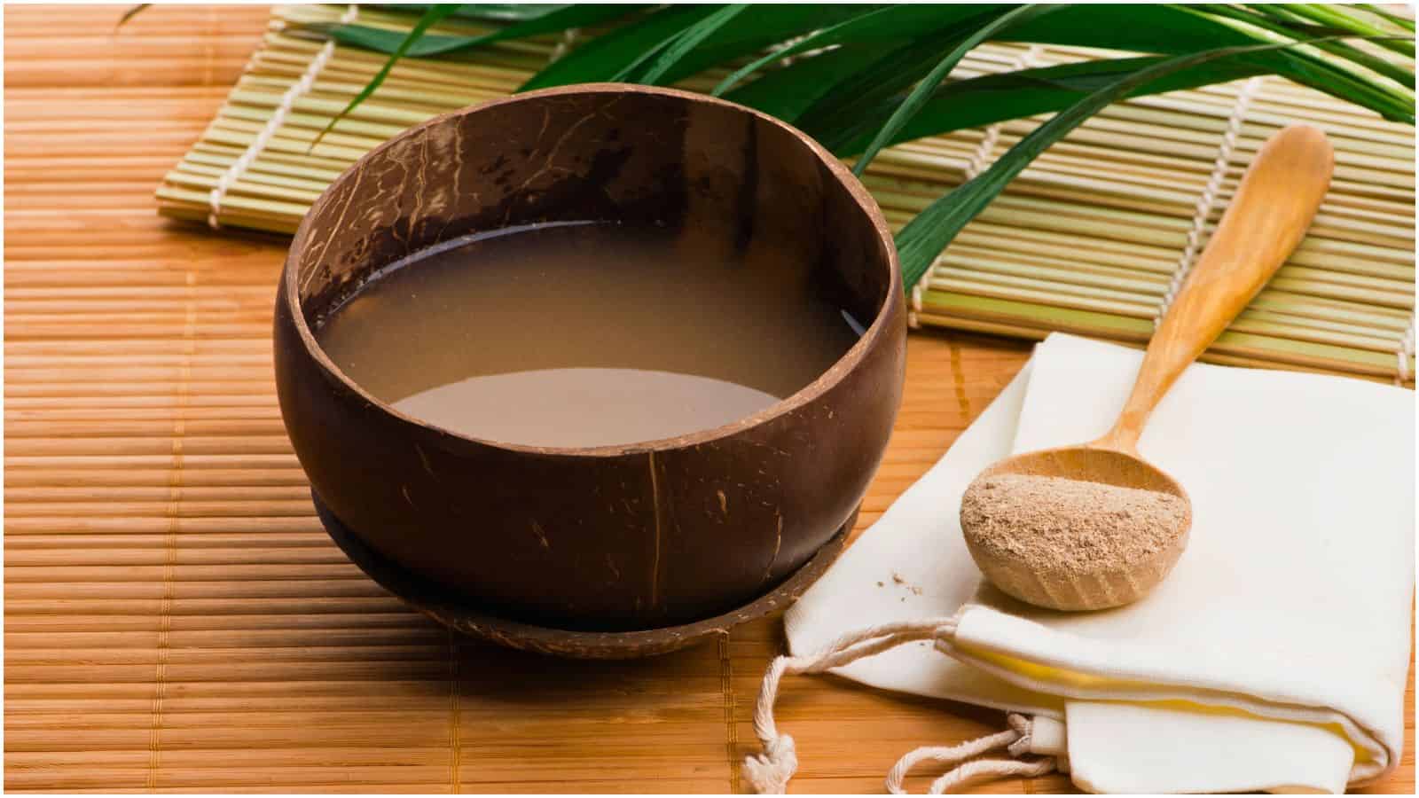 A coconut shell bowl filled with a brown liquid is placed on a bamboo mat. A wooden spoon with a pile of brown powder rests on a folded cloth next to it. Green leaves and a rolled bamboo mat are in the background.